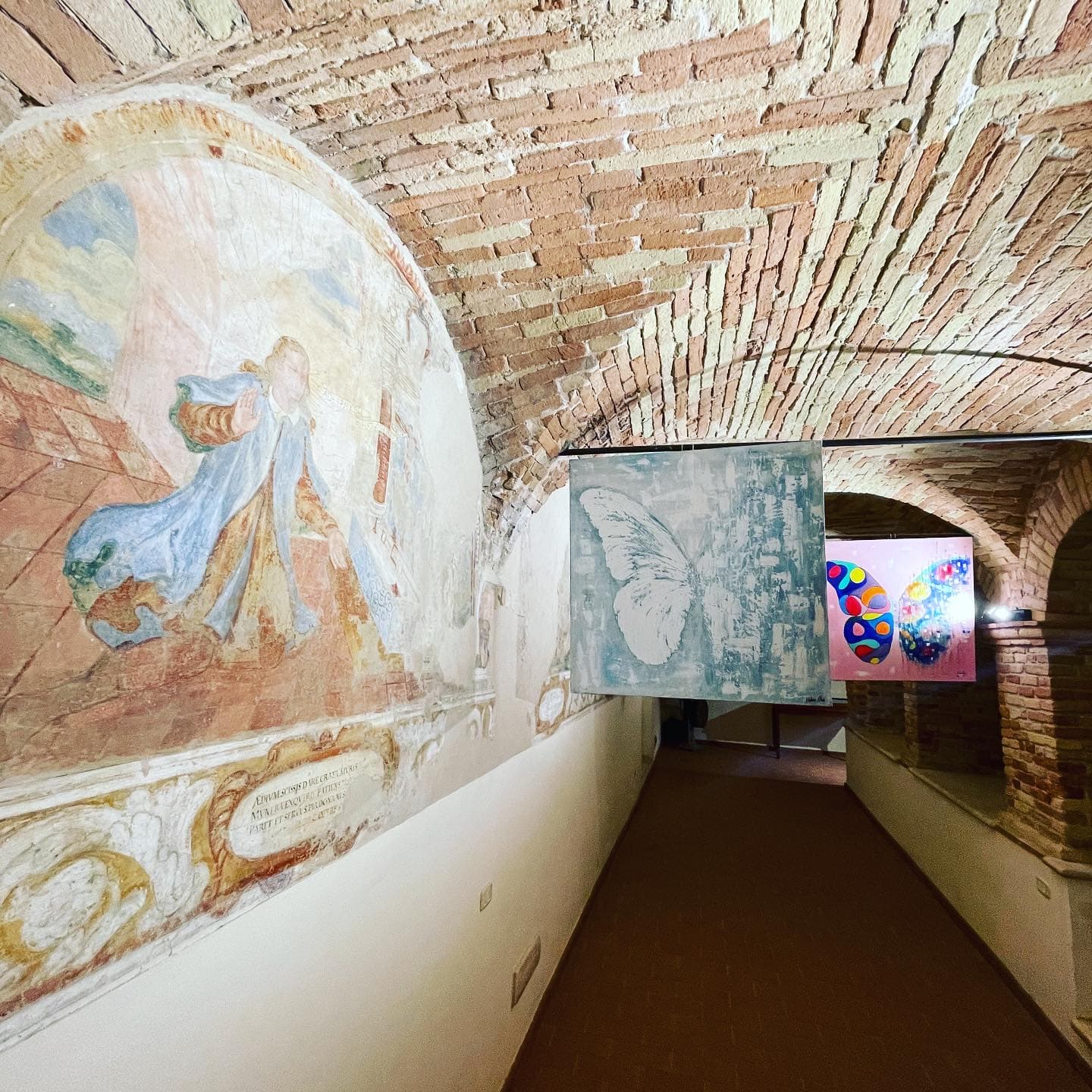 A vaulted brick corridor with frescoes; butterfly paintings hang along the passage.