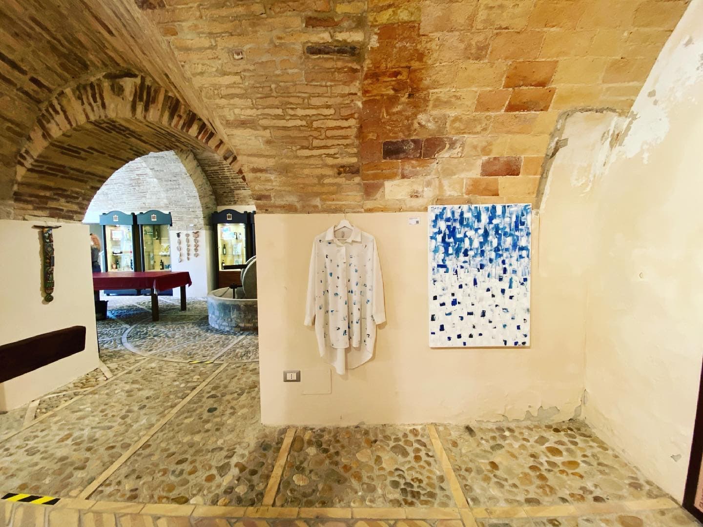 A vaulted brick hall with a cobbled floor; a blue and white painting hangs on the wall.