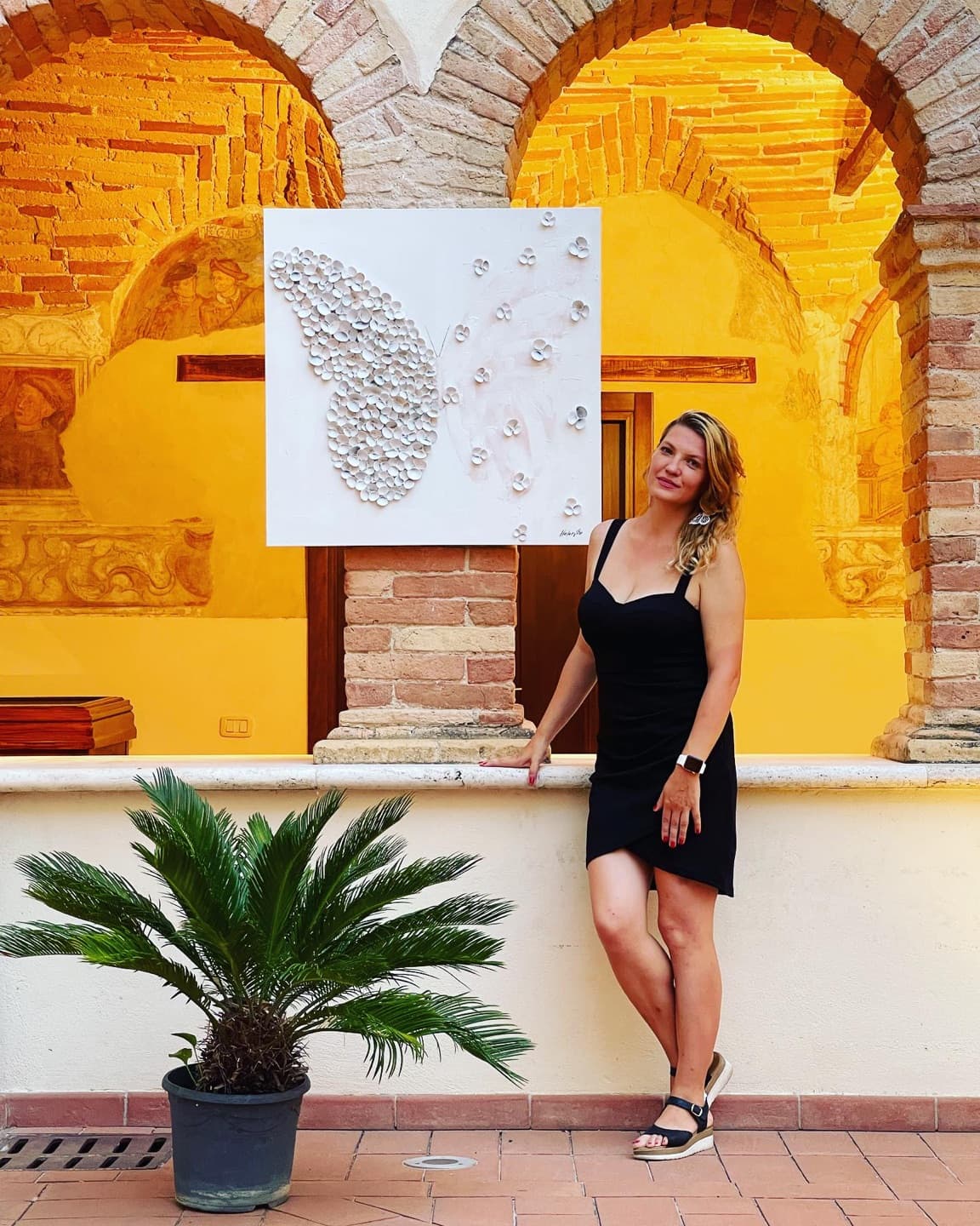 Helen She in the lit cloister arcade in the evening, beside a white butterfly work.