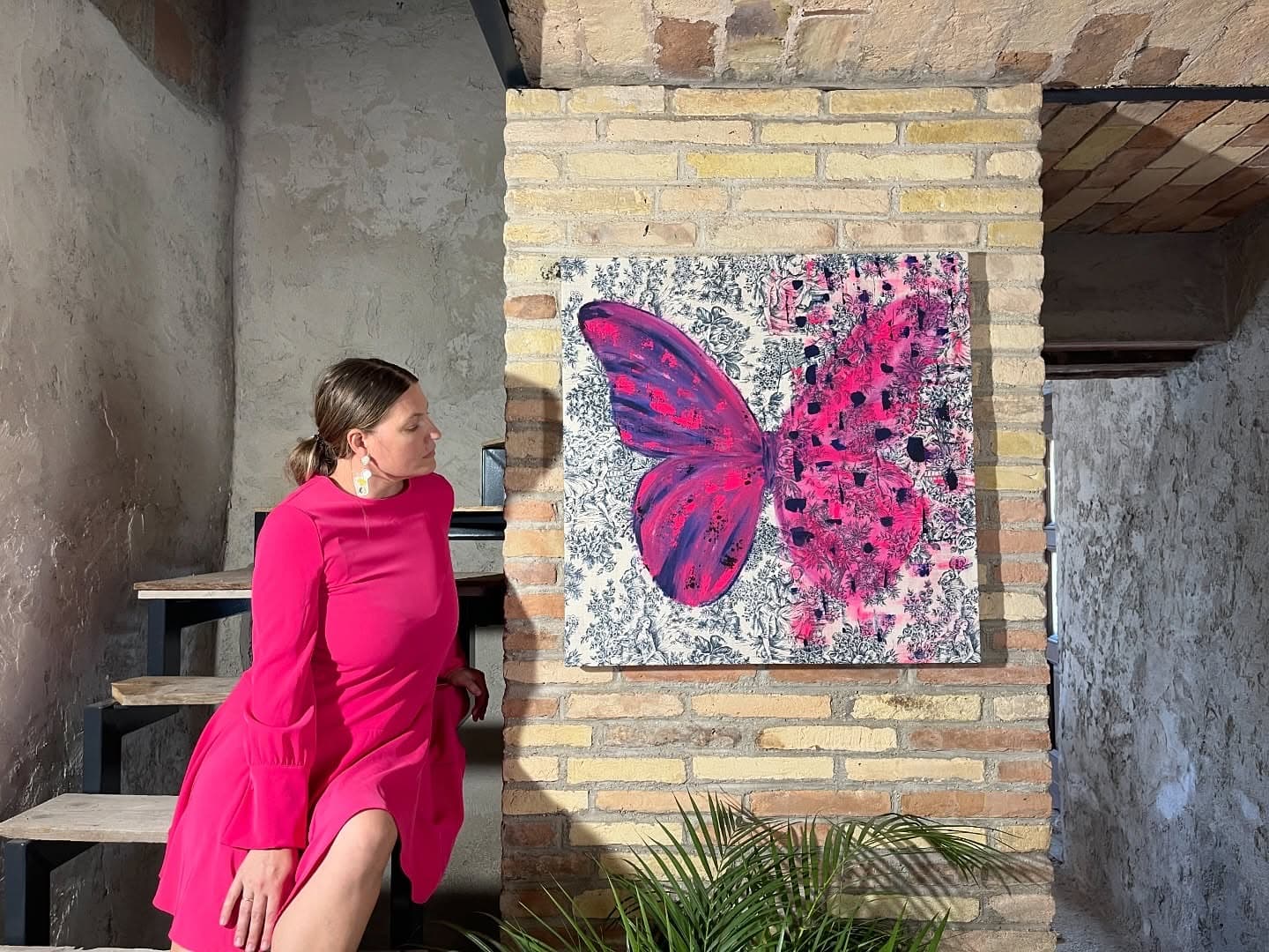 Helen She in a pink dress beside a pink and black butterfly painting hung on a brick wall.