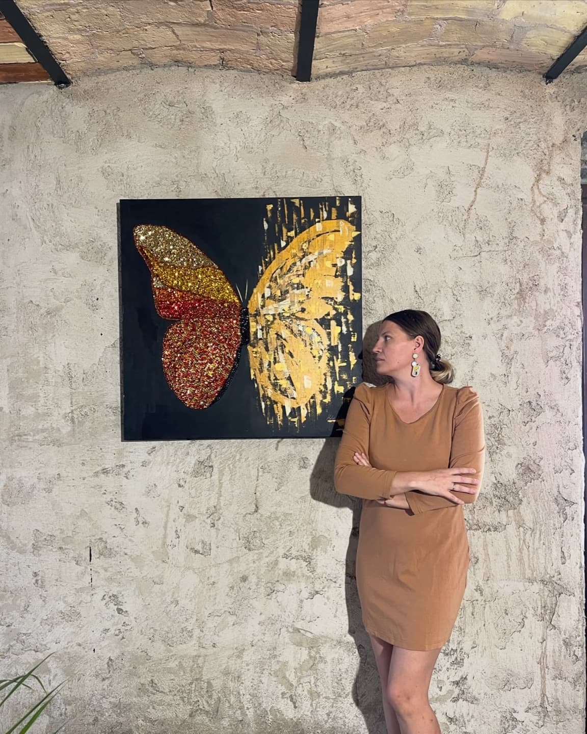 Helen She leaning against a plaster wall next to a red and gold butterfly painting on a black ground.