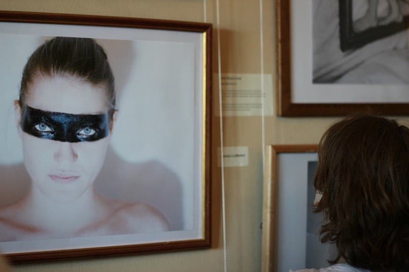 A framed photograph of Helen with a dark painted band across her eyes, hanging in a museum hall beside other framed works.